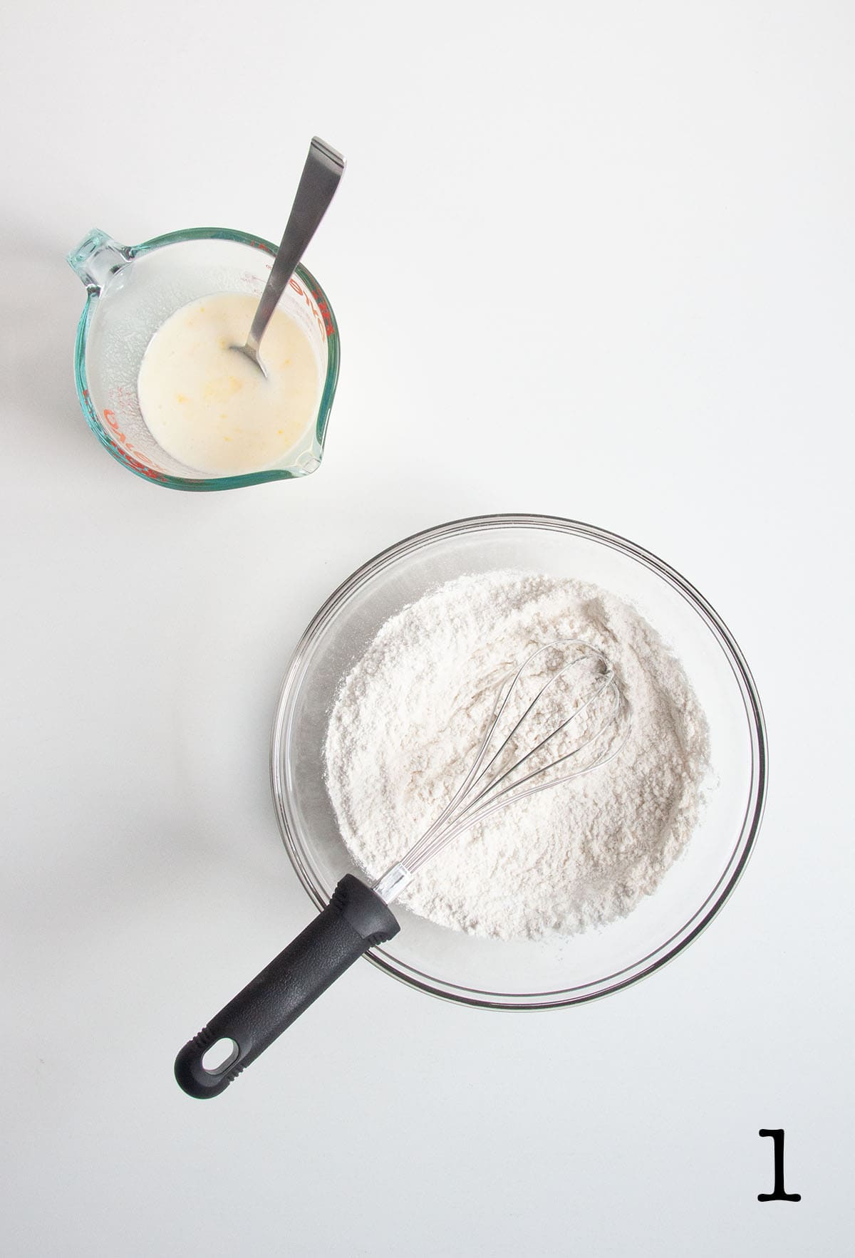 A bowl of dry pancake mix with a whisk resting inside and a glass beaker with wet ingredients next to it.