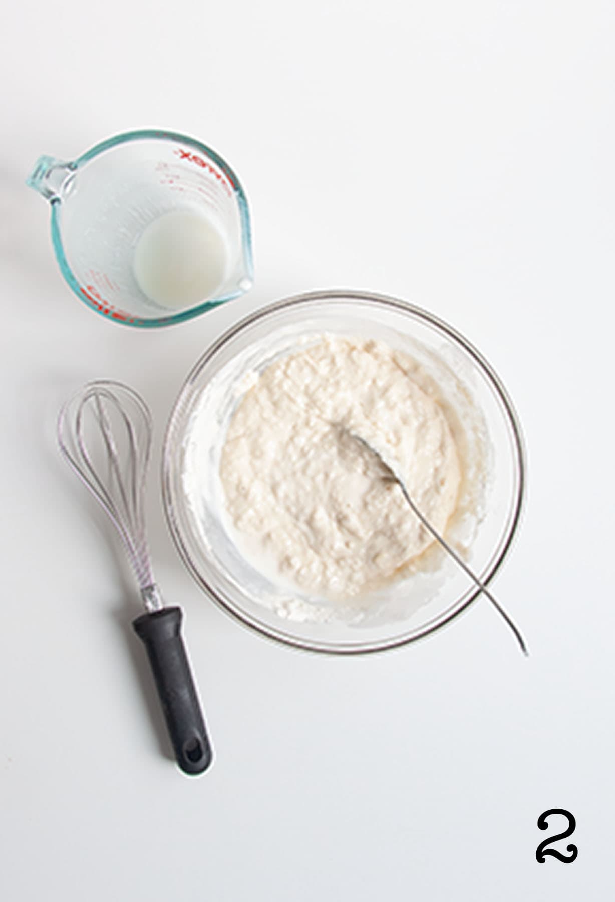 A glass bowl filled with pancake batter, and a whisk and an empty liquid measuring cup beside it.