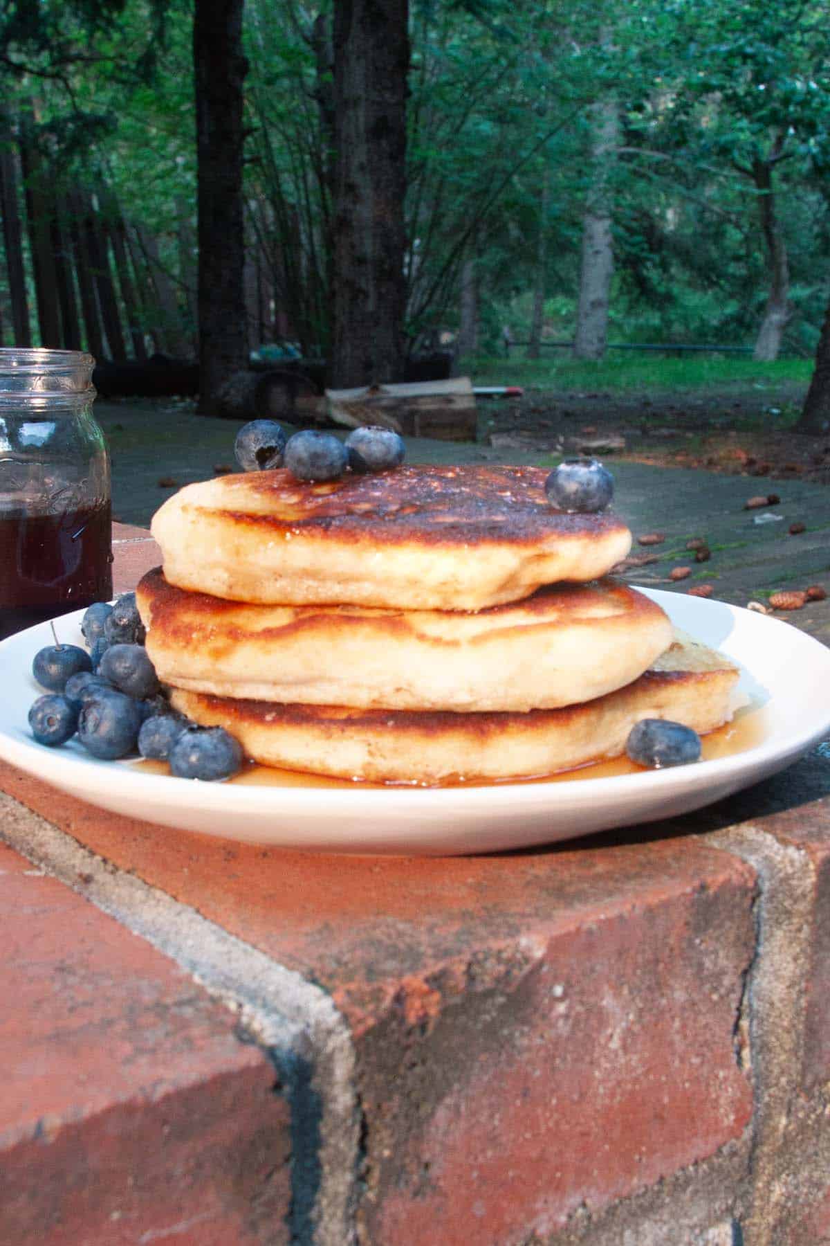 A stack of pancakes, maple syrup, and blueberries on a plate outside.