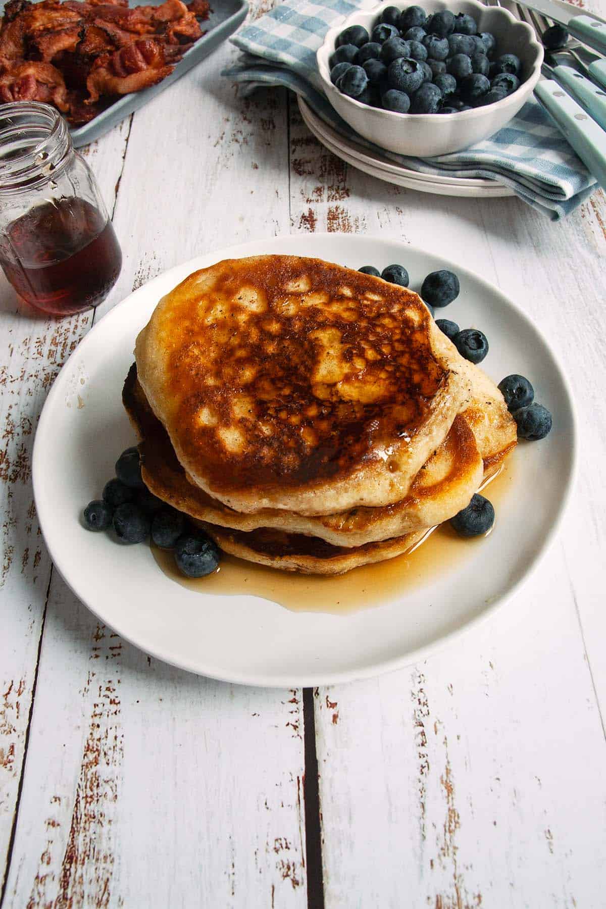 A stack of pancakes, maple syrup, and blueberries on a plate with blueberries and bacon in the background.