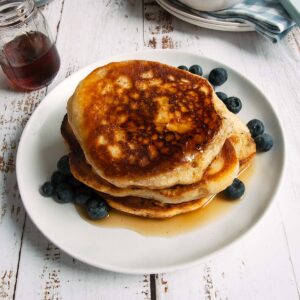 A stack of pancakes, maple syrup, and blueberries on a plate with blueberries and bacon in the background.