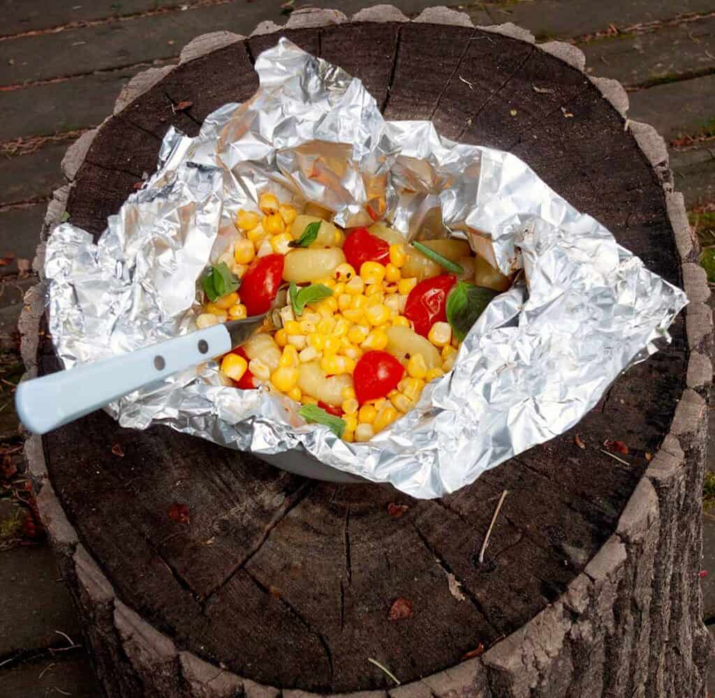 An open foil packet filled with gnocchi, corn, tomatoes, and basil on a wooden tree stump.