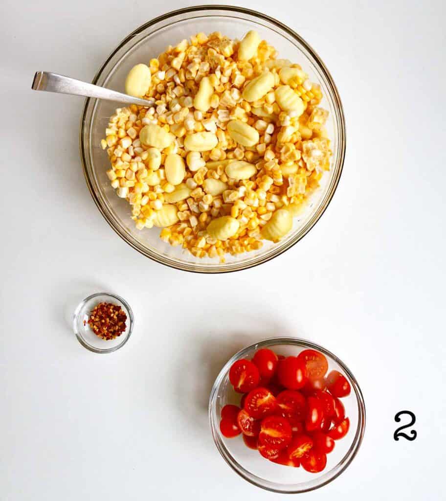 A bowl of corn and gnocchi, with bowls of tomatoes and pepper flakes nearby.