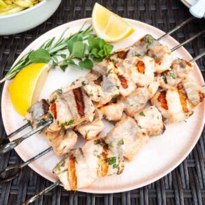 Four grilled salmon kabobs on a pink plate with lemon wedges and fresh herbs next to them.