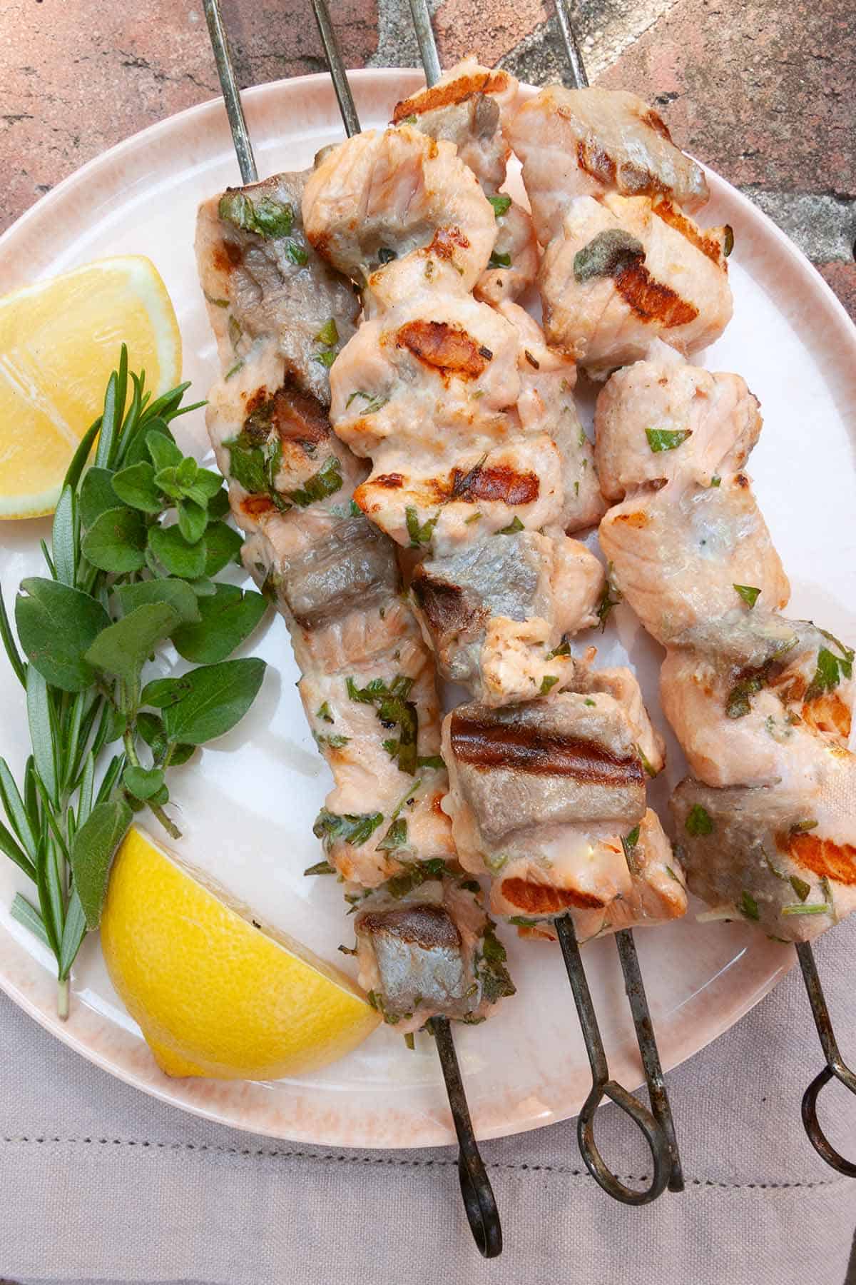 Four grilled salmon skewers on a pink plate with fresh herbs and lemon wedges.