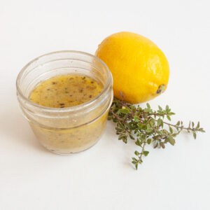 A jar of lemon thyme vinaigrette wit ha lemon and bunch of thyme behind it.