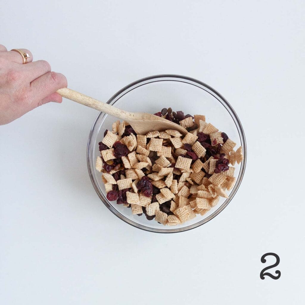A person stirring a bowl of trail mix with Chex, dried cranberries, pepitas, and chocolate chips.