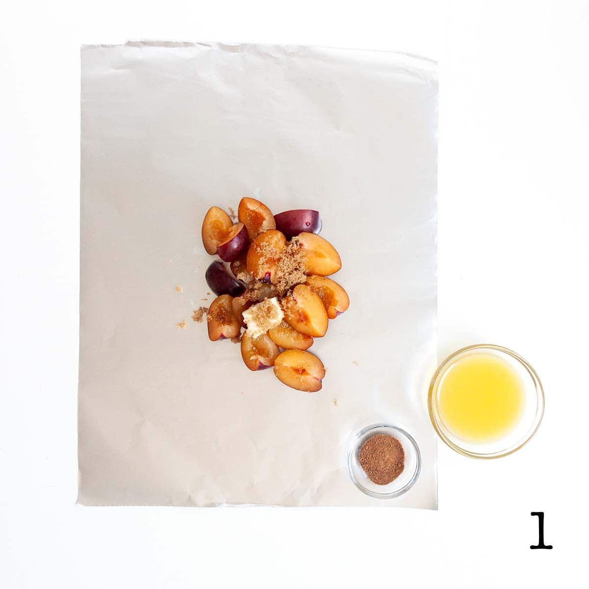 Quartered plums with butter and brown sugar on a foil sheet with bowls of nutmeg and orange juice on the side.