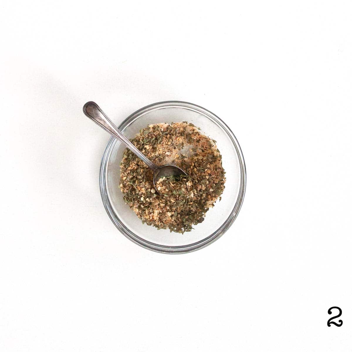 A glass bowl with spices mixed together and spoon resting inside.