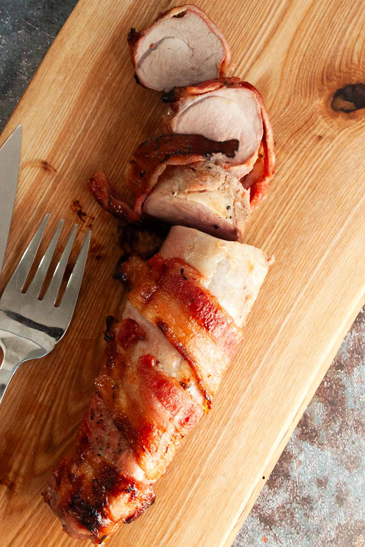 A partially sliced grilled bacon-wrapped pork tenderloin on a wooden cutting board.