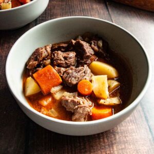 A bowl of beef stew with carrots and potatoes on a wooden table.