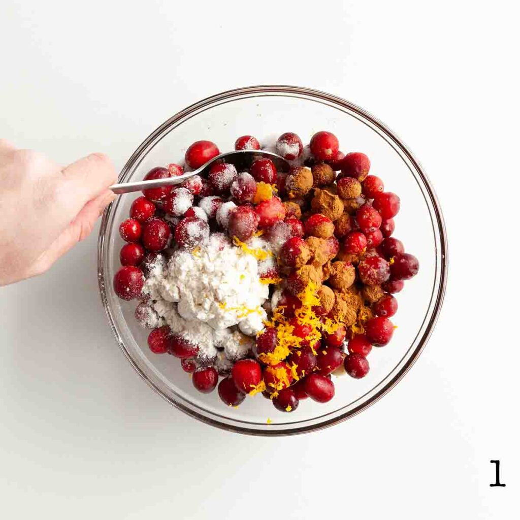 A person mixing cranberries, orange zest, flour, sugar, and cinnamon in a glass bowl.