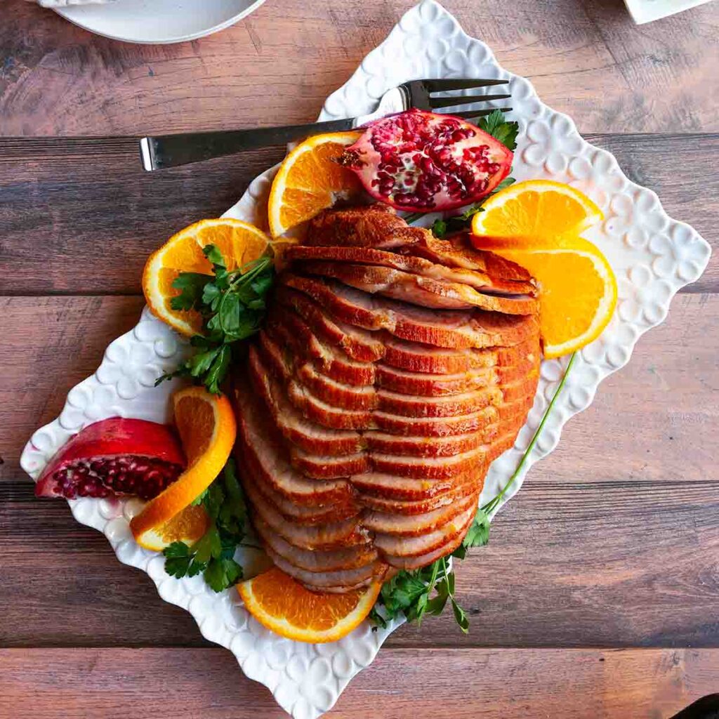 A spiral cut ham on a rectangular platter with orange slices, pomegranate wedges, and parsley sprigs.