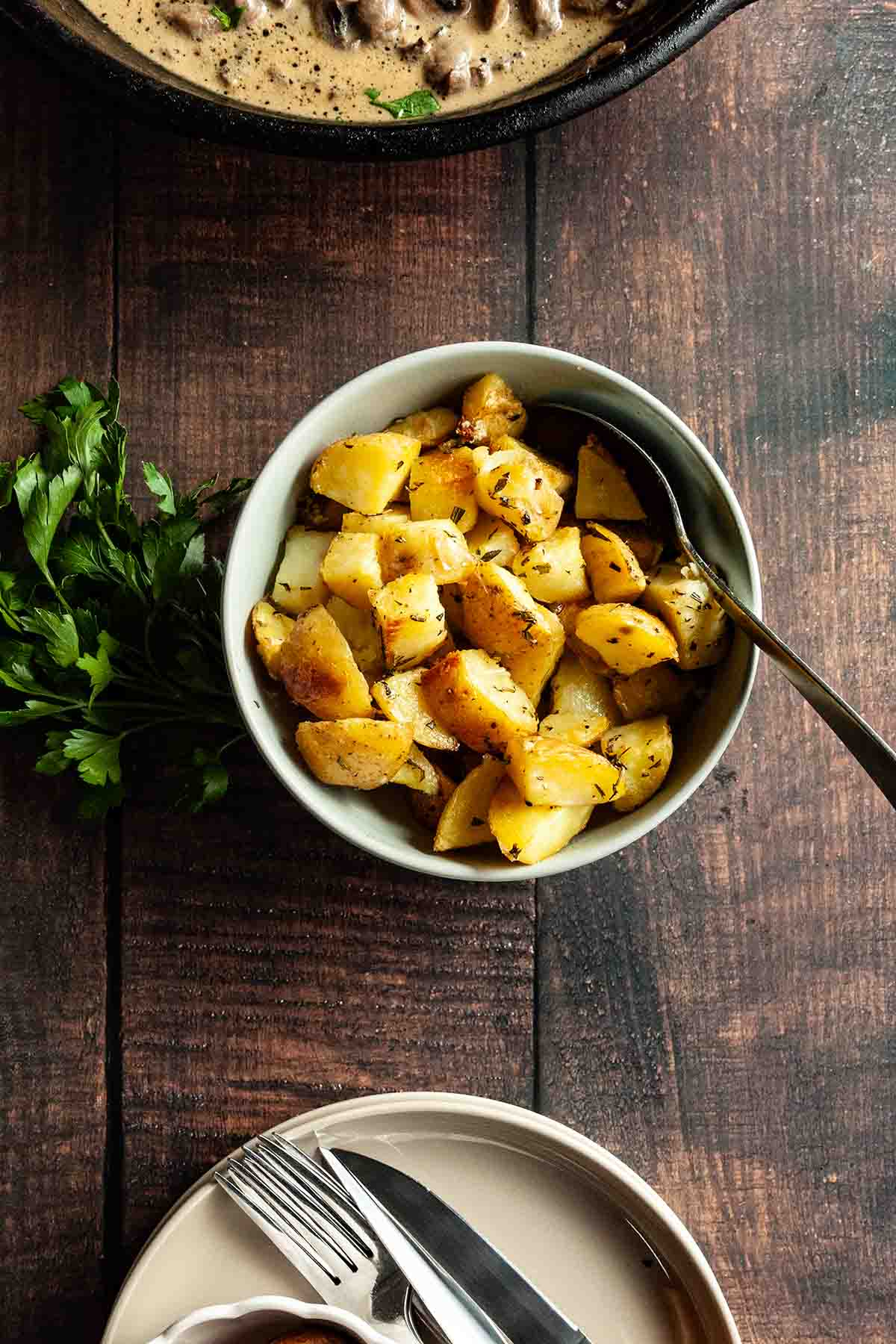 A bowl of roasted potatoes on a table with plates and utensils, fresh parsley, and a skillet meal.