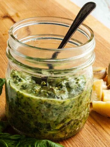 A jar of nut-free pesto on a cutting board with fresh basil, garlic, and parmesan on the side.