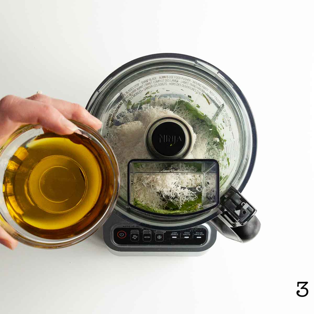 A person pouring oil into a food processor filled with pesto ingredients.