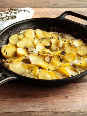 A cast iron skillet filled with scalloped potatoes with sprigs of thyme on top.
