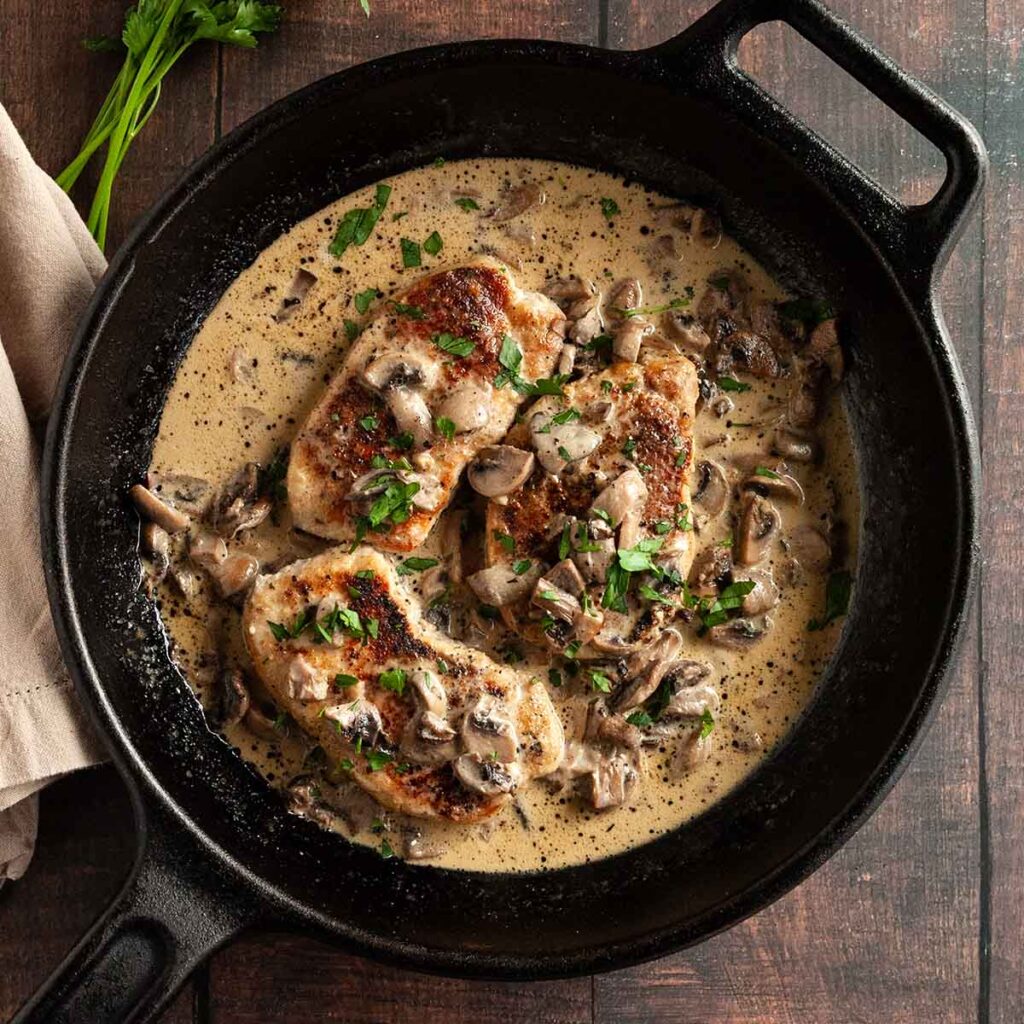 Three pork chops in a cast-iron skillet with mushroom gravy on top and around the pork chops.