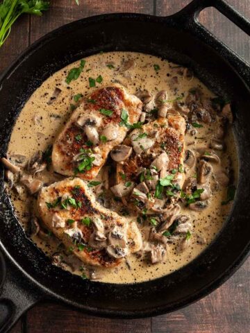 Three pork chops in a cast-iron skillet with mushroom gravy on top and around the pork chops.
