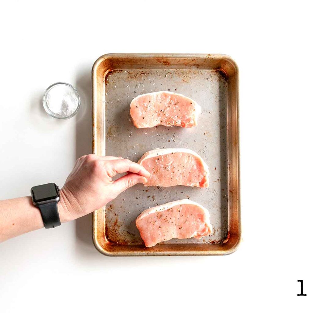 A person seasoning three pork chops with salt on a baking sheet.