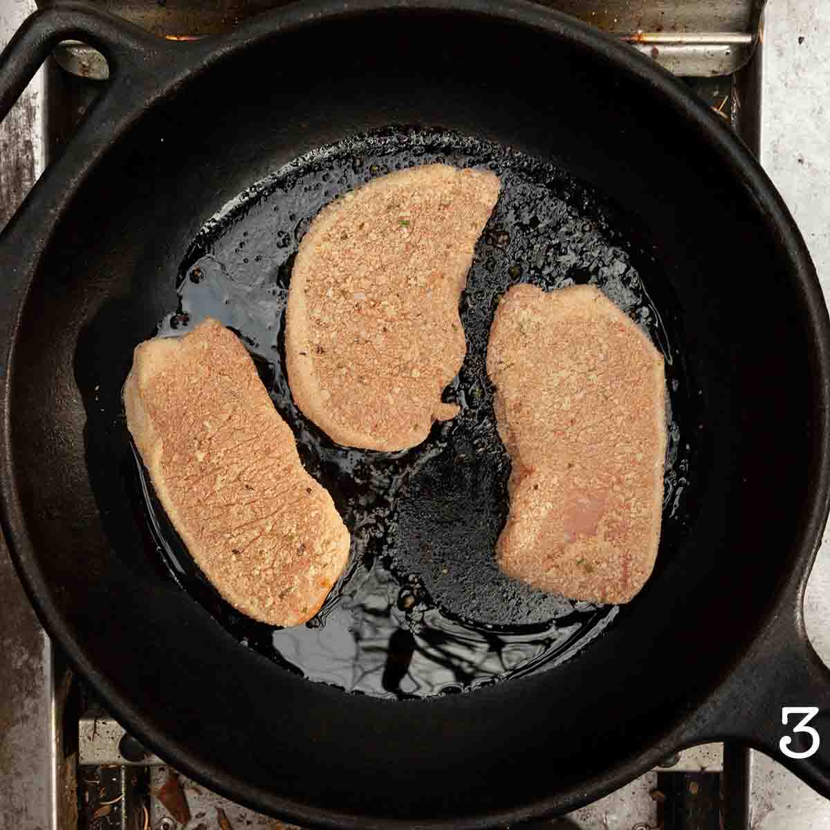 Three pork chops coated with fine bread crumbs in a cast iron skillet.