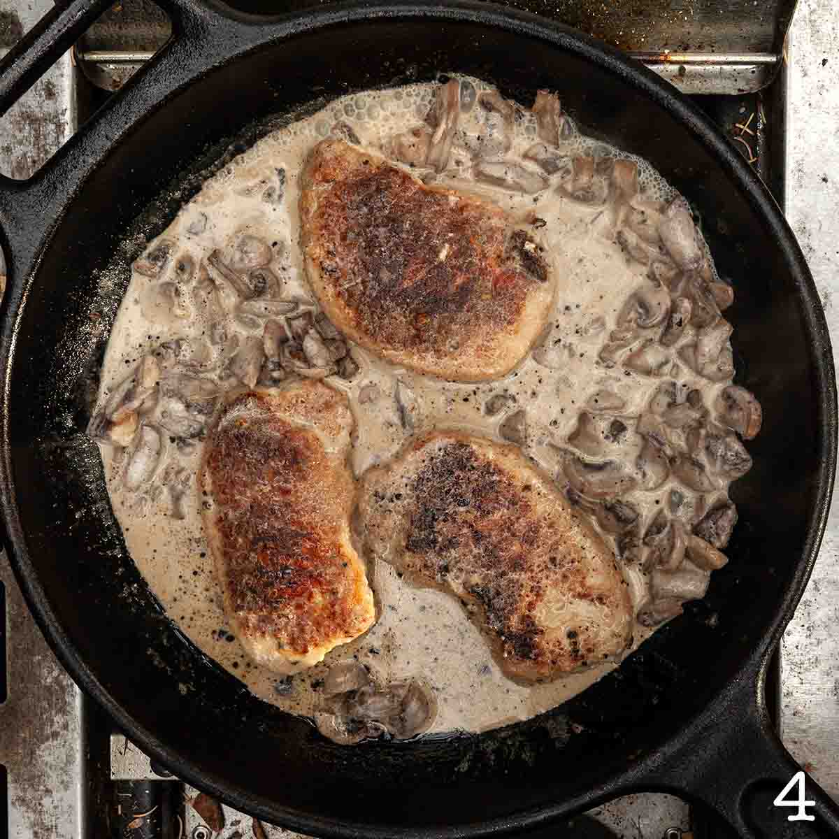 Three cooked breaded pork chops in a cast iron skillet with mushroom gravy simmering around them.