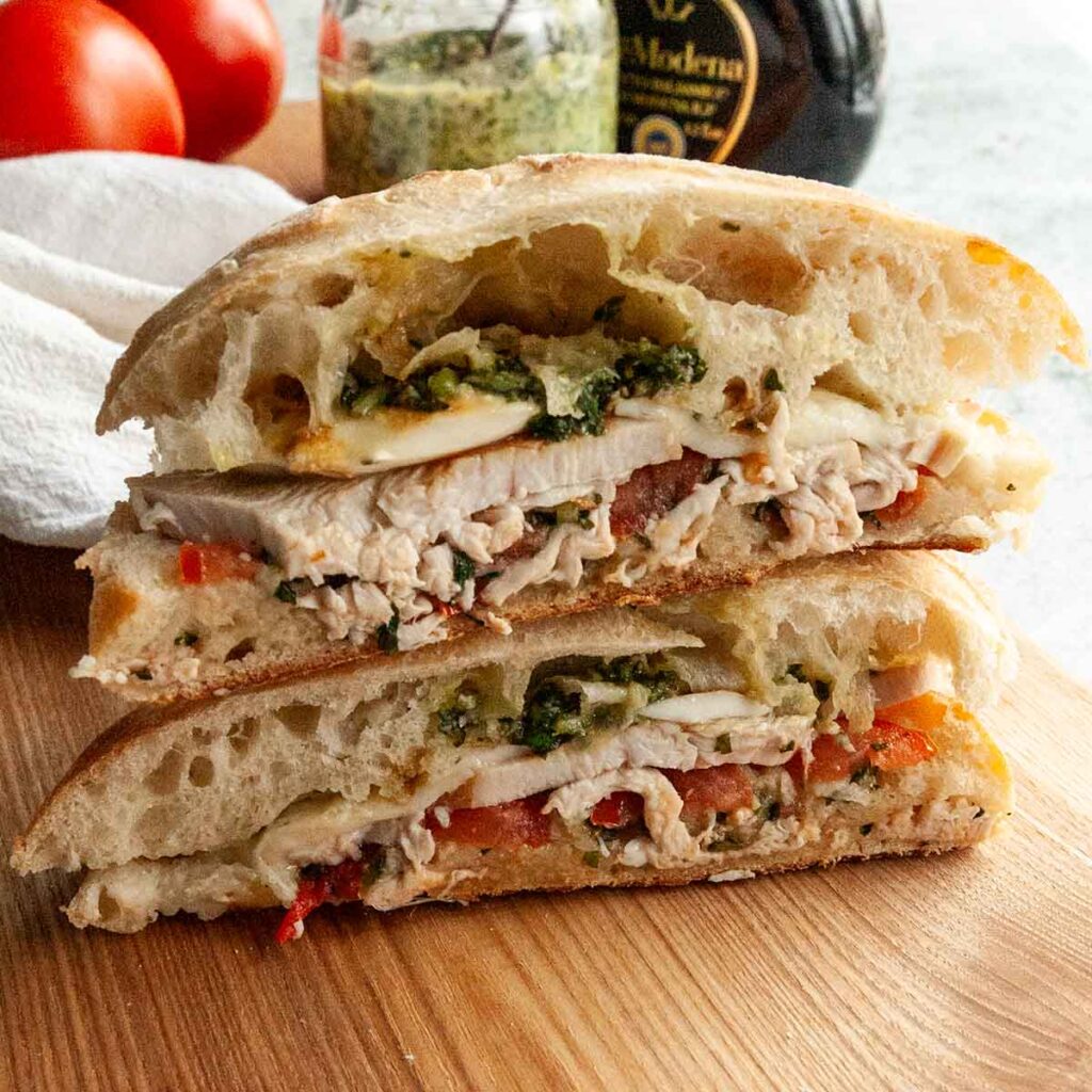 Two halves of a turkey ciabatta sandwich with tomatoes, pesto, and mozzarella, stacked on top of each other on a cutting board.