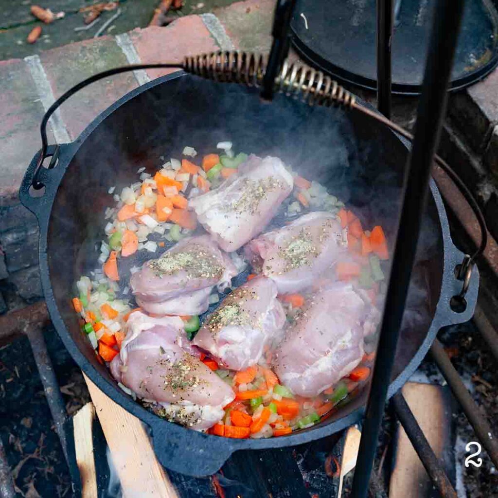 Seasoned chicken thighs on top of sautéed vegetables in a Dutch oven over a fire.