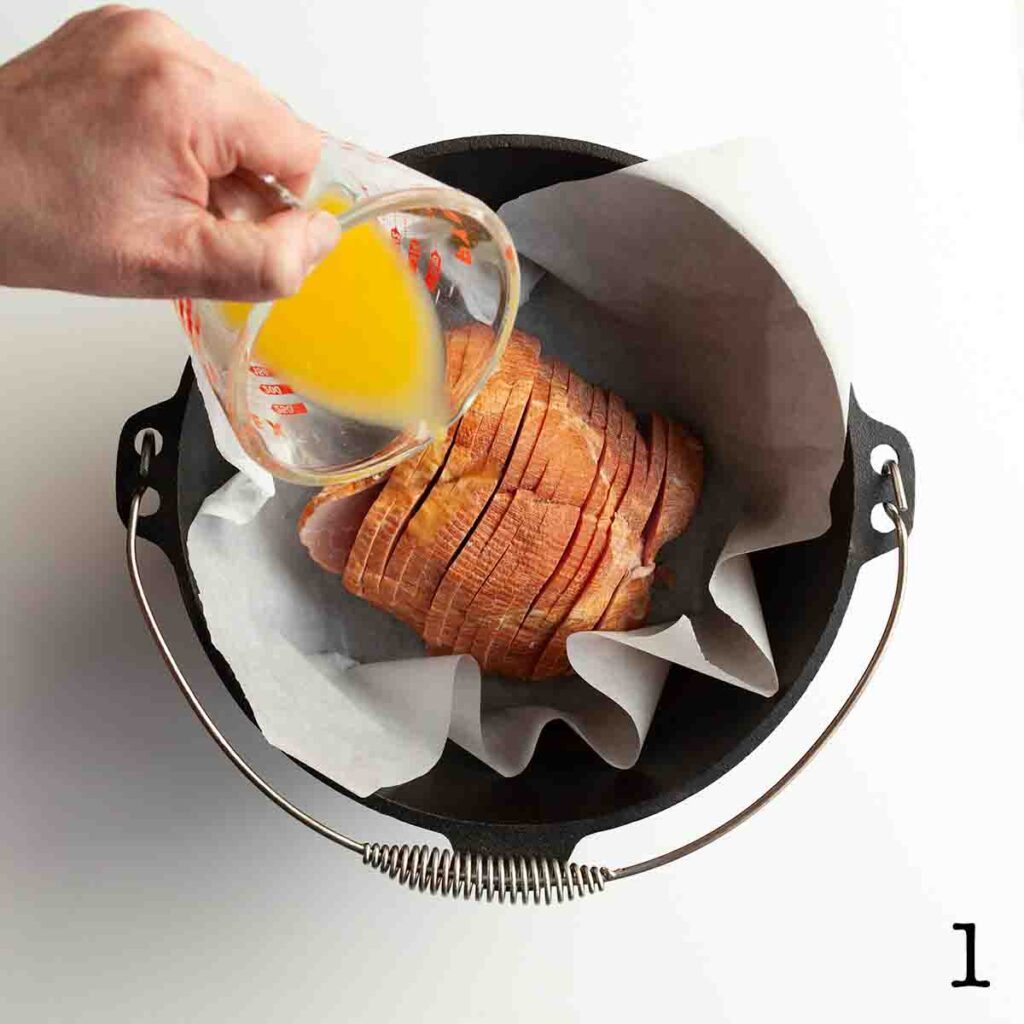A person pouring orange juice over a spiral cut ham in a Dutch oven.