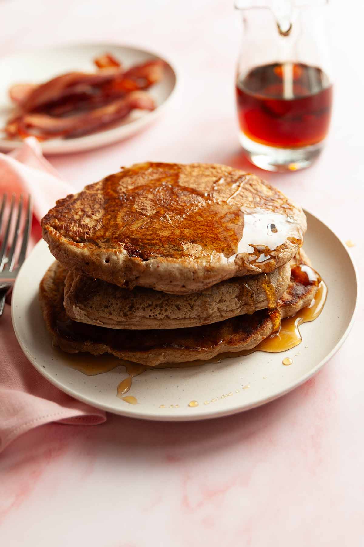 A stack of gingerbread pancakes on a plate with a pitcher of maple syrup and a plate of bacon in the background.