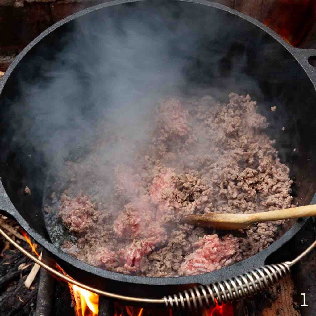 Meat being browned in a cast iron Dutch oven over a fire.