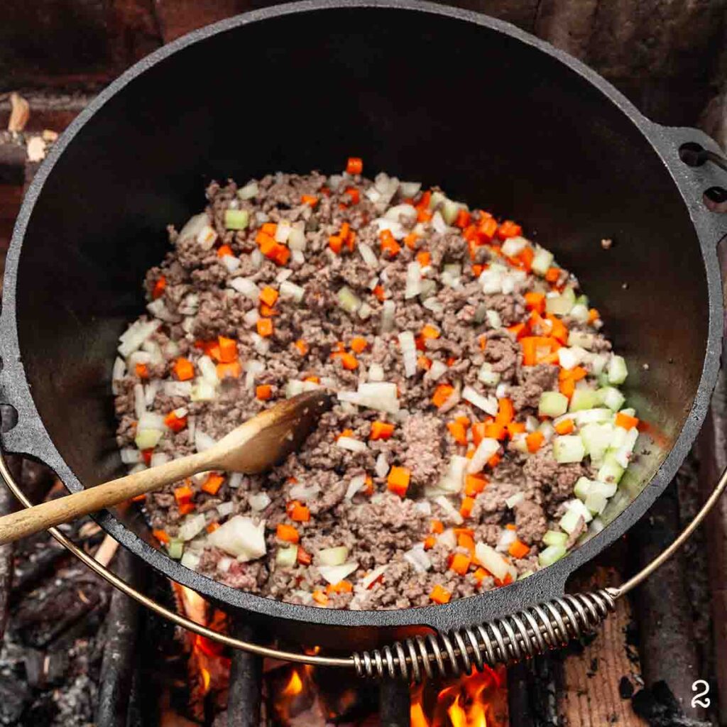 Raw carrot, onion, and celery in a Dutch oven with cooked ground meat over a fire.