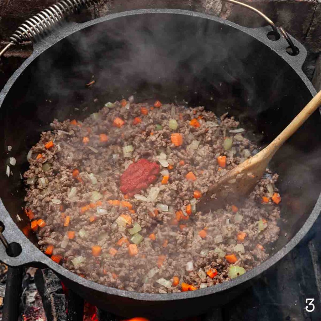 A Dutch oven filled with ground beef, onion, carrots, and celery with a dollop of tomato paste in the center.