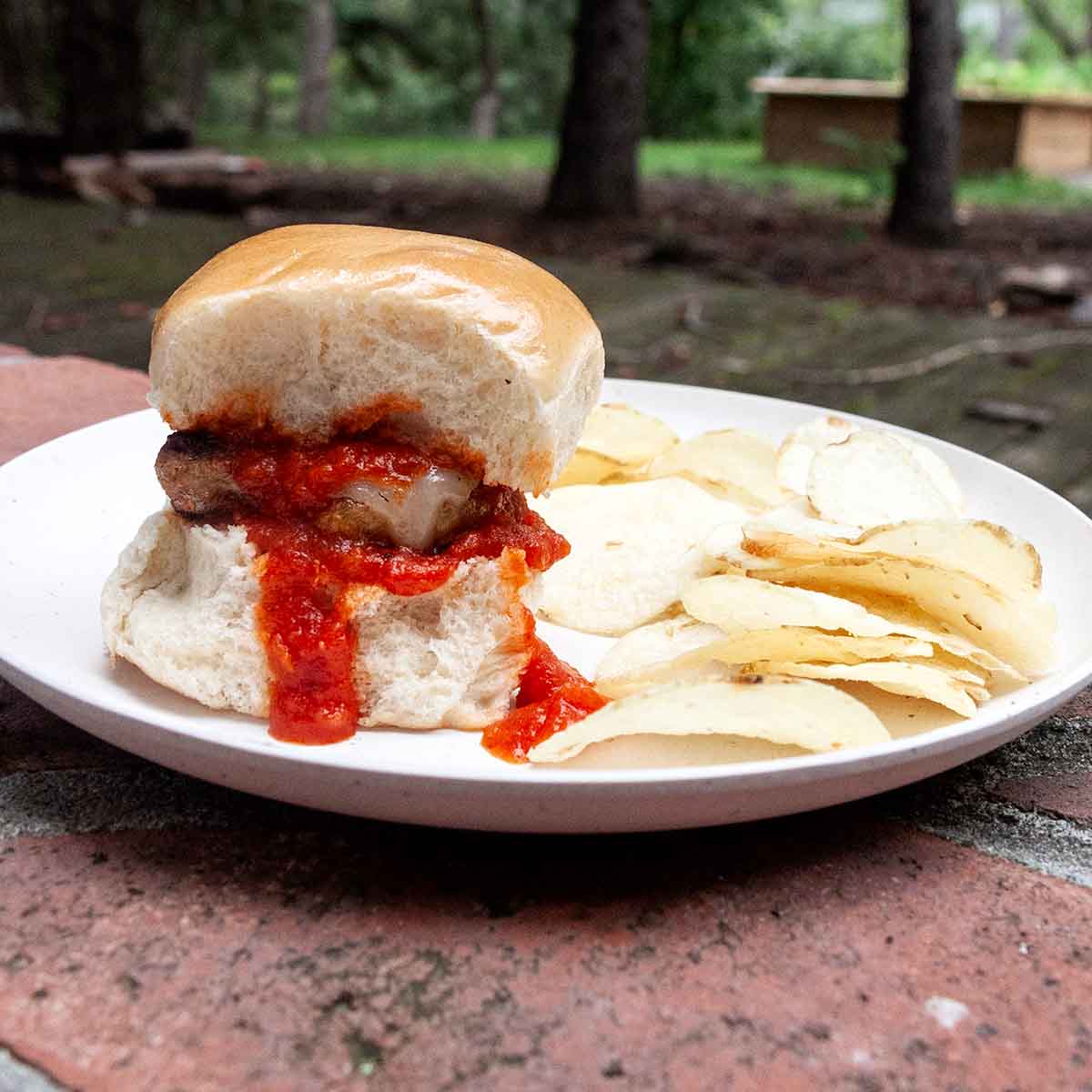 An Italian sausage slider topped with marinara on a plate with potato chips on the side.