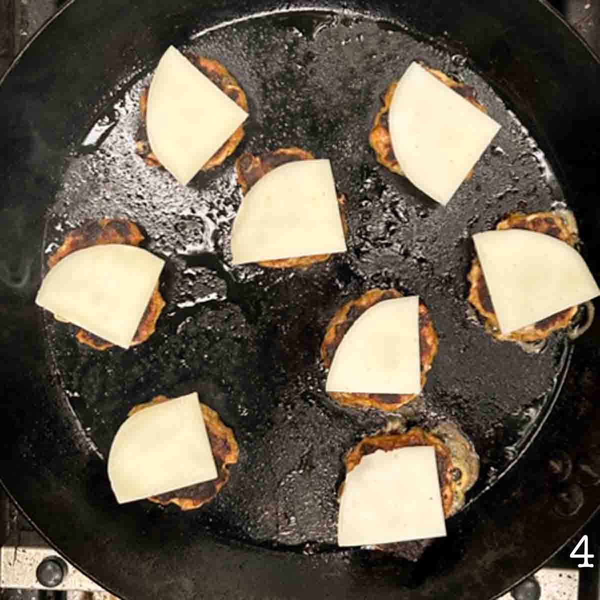 Eight sausage slider patties in a skillet with pieces of provolone melting on top.