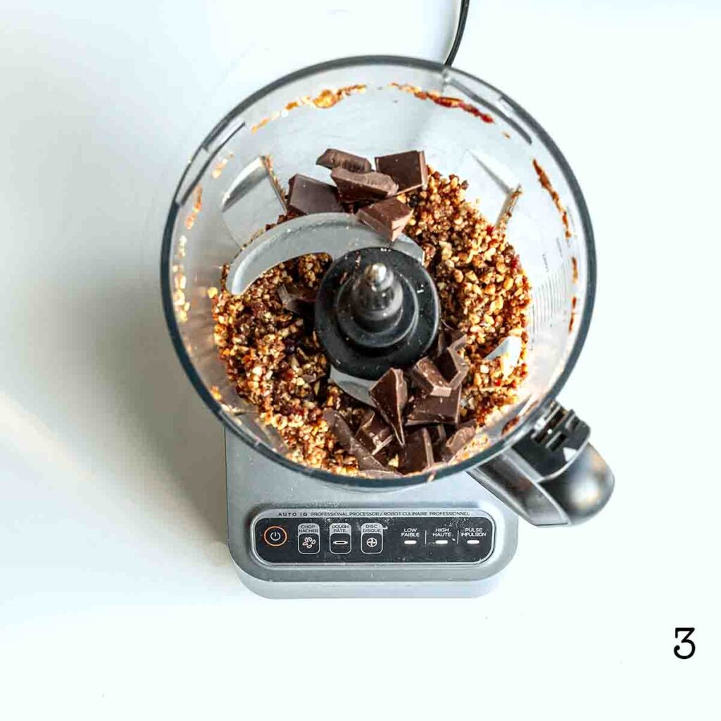 A food processor with ground nuts and fruit and pieces of dark chocolate.