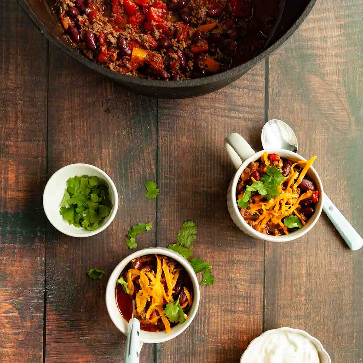 A pot of camping chili with two mugs of chili next to it, topped with cheese and cilantro.