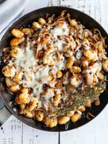 A cast iron skillet filled with French onion gnocchi with a bunch of thyme on the side.