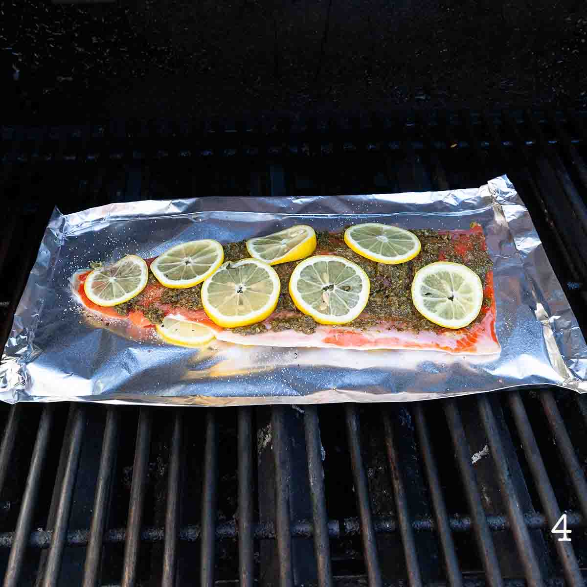 A fillet of salmon with pesto and lemon slices on top on a sheet of foil on a grill.