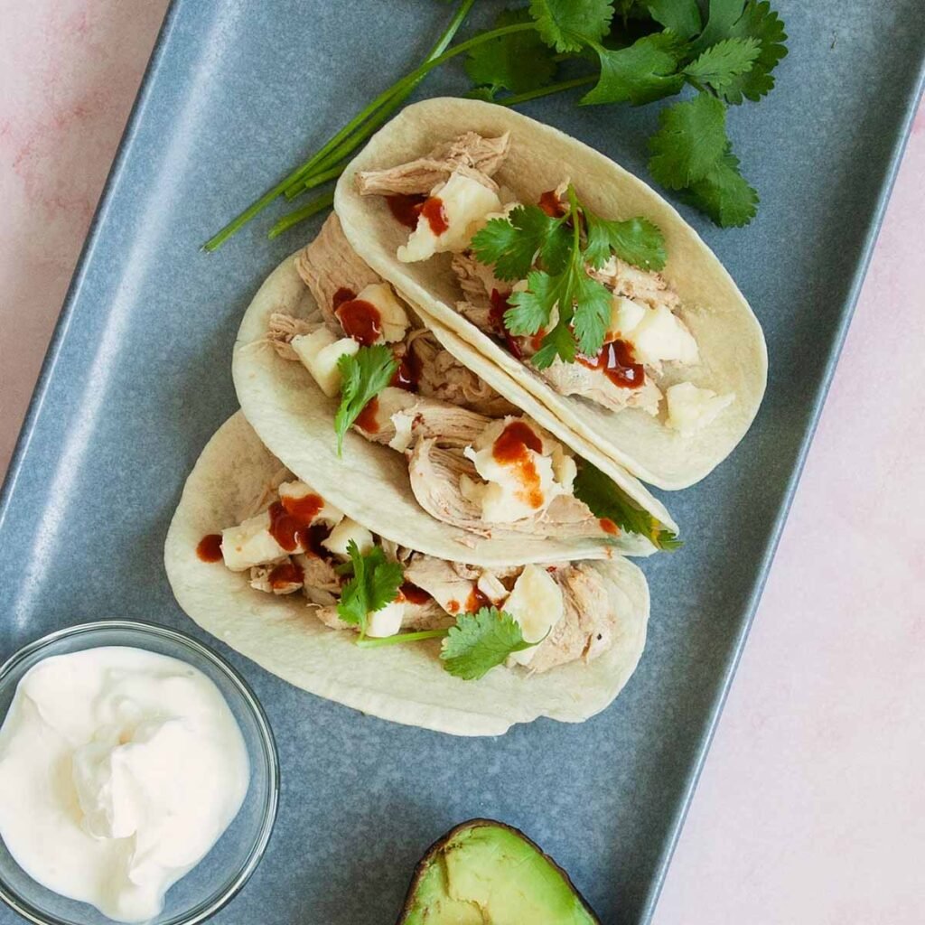 Three shredded chicken tacos on a blue platter with sour cream, avocado, and cilantro.