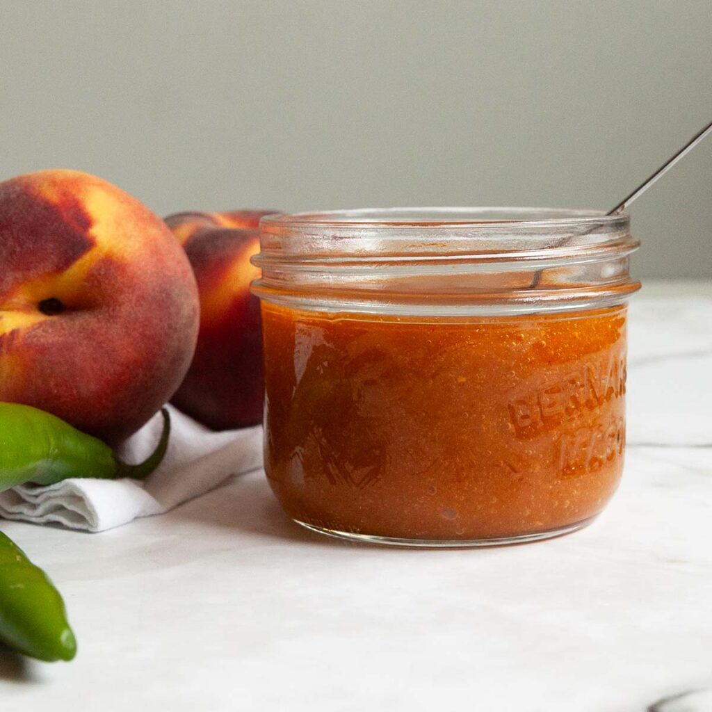 A small jar filled with peach bbq sauce with two peaches and two serrano peppers next to it.