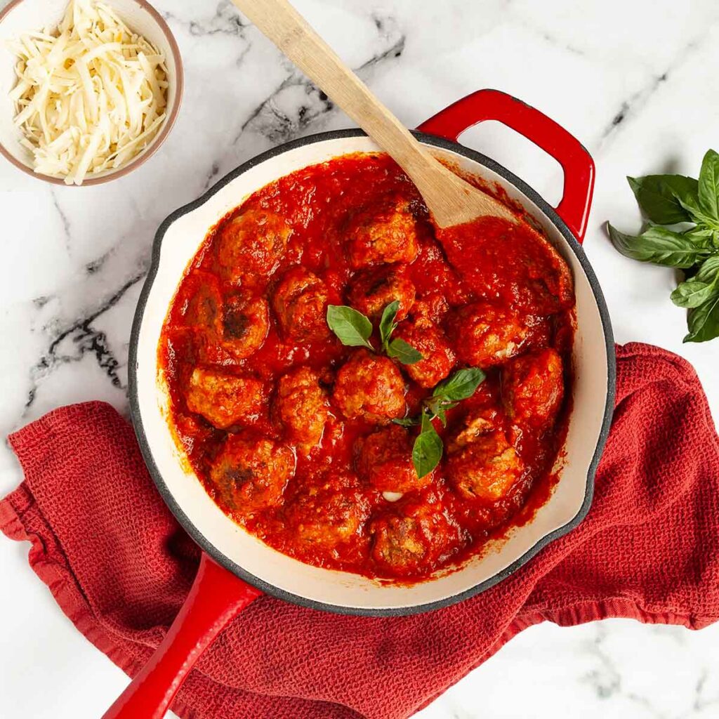 A skillet filled with sausage meatballs in a marinara sauce with shredded cheese on the side.