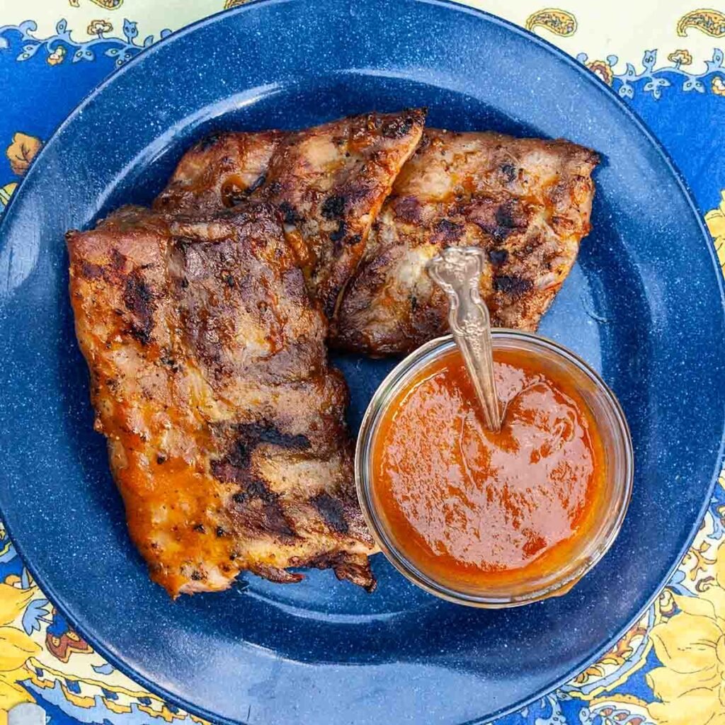 Three pieces of grilled baby back ribs on a blue plate with a jar of bbq sauce.