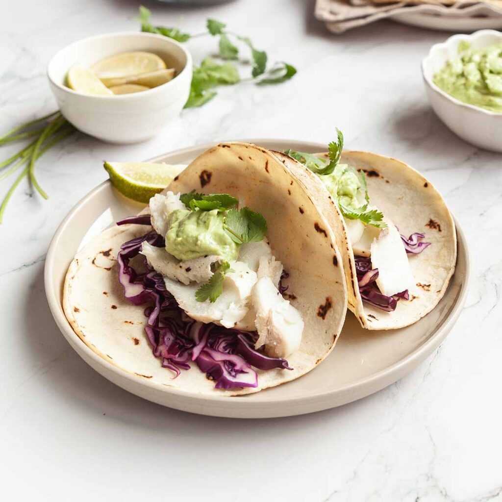 Two halibut tacos on a plate, with red cabbage, avocado crema, and cilantro on top.