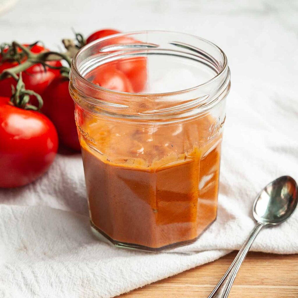 A jar of sun-dried tomato vinaigrette with tomatoes in the background.