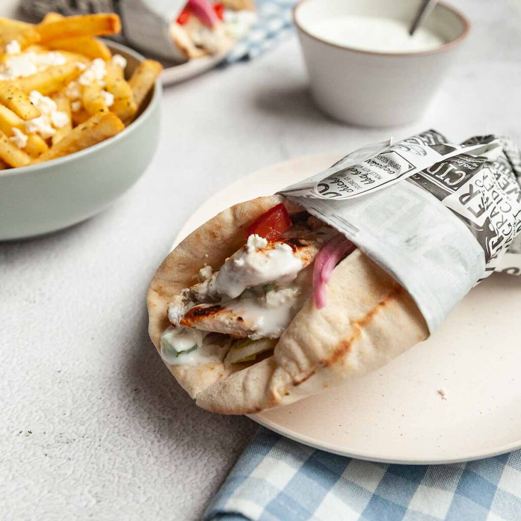 A grilled chicken gyro on a plate with a bowl of fries and a bowl of dip in the background.