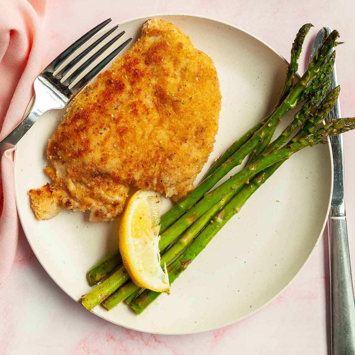 A breaded chicken cutlet on a plate with some asparagus and a lemon wedge.