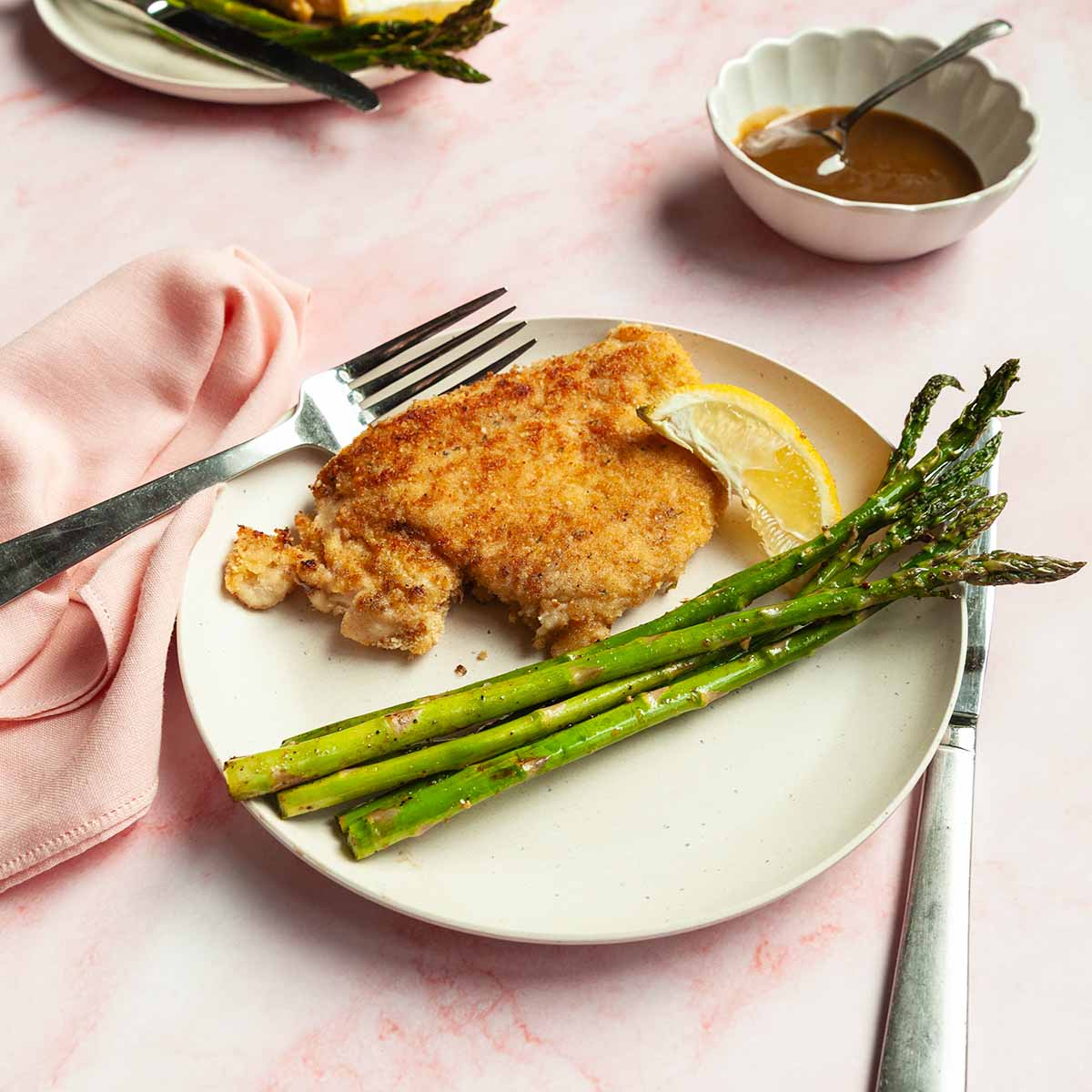 A breaded chicken cutlet on a plate with some asparagus and a lemon wedge.