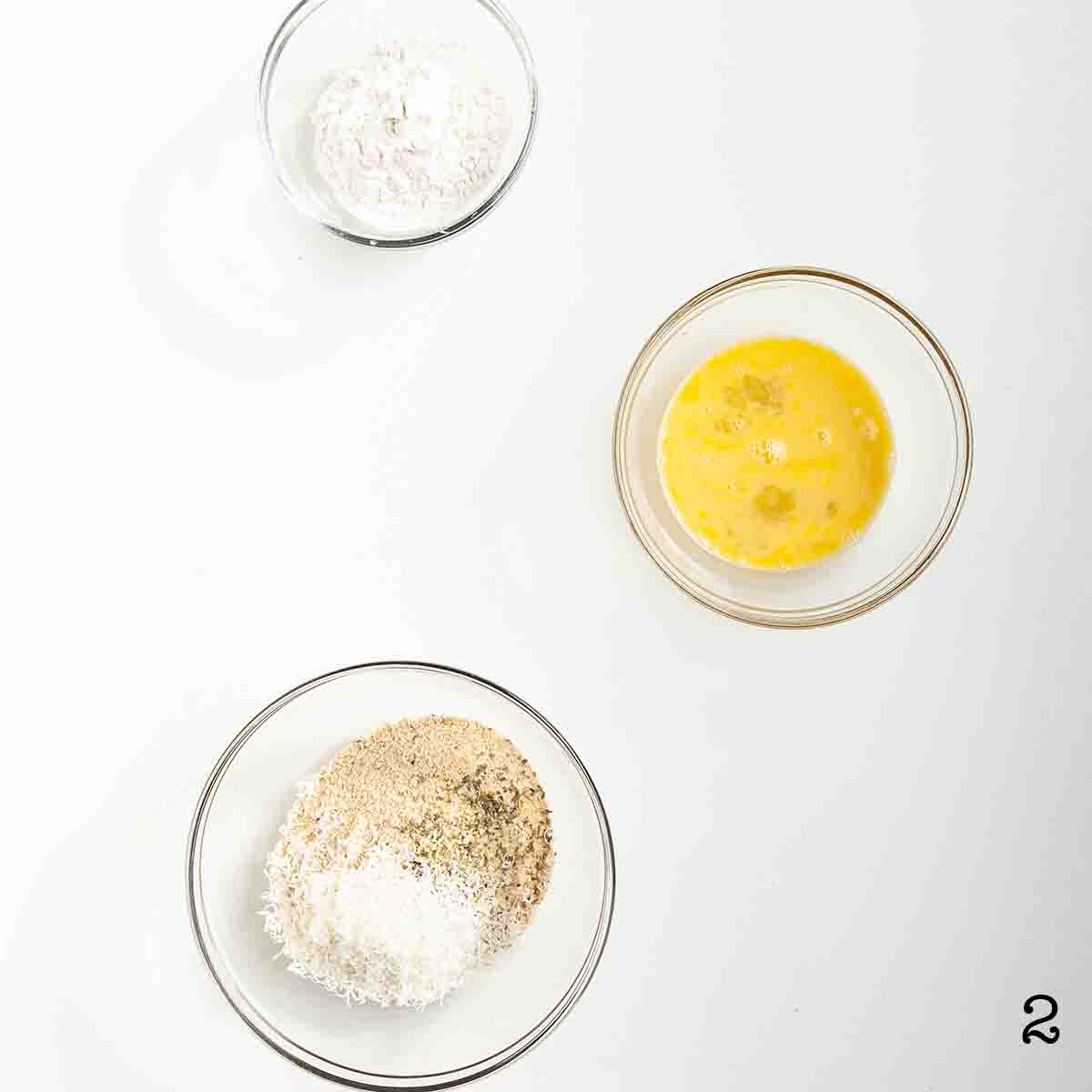 A bowl of flour, a bowl of beaten egg, and a bowl of bread crumbs with Parmesan.