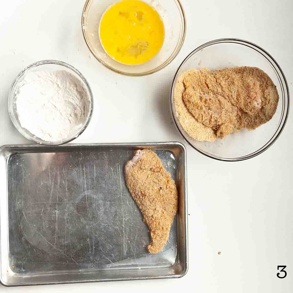 A breaded chicken cutlet on a baking sheet with breading bowls nearby.